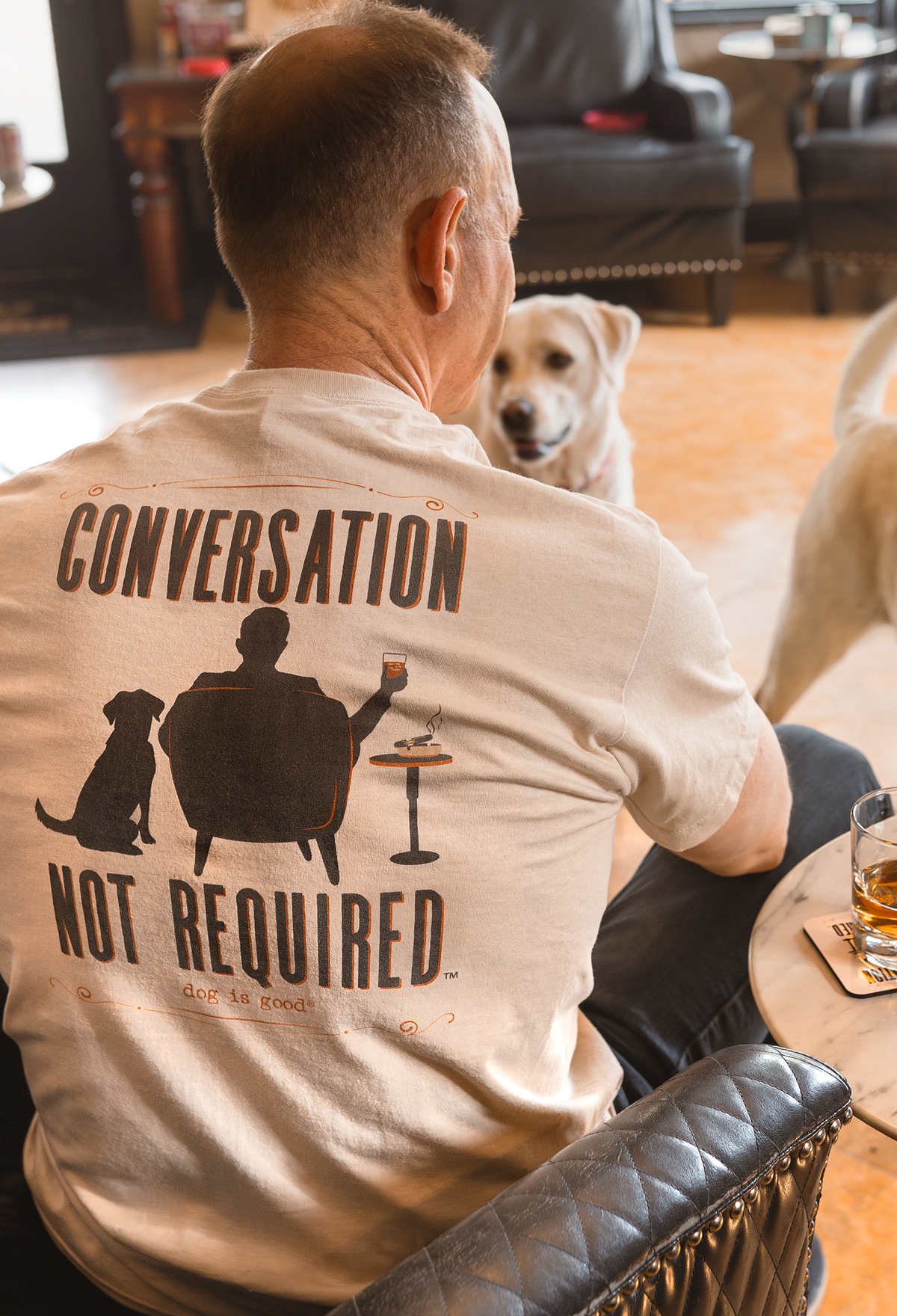 Man in a beige shirt with a dog graphic, sitting near two dogs and a table with drinks.