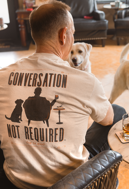 Man in a beige shirt with a dog graphic, sitting near two dogs and a table with drinks.