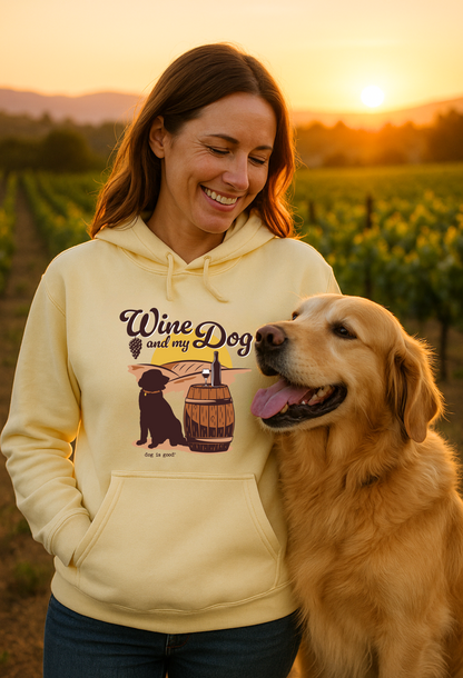 Woman in a yellow hoodie smiles next to a golden retriever in a vineyard at sunset.